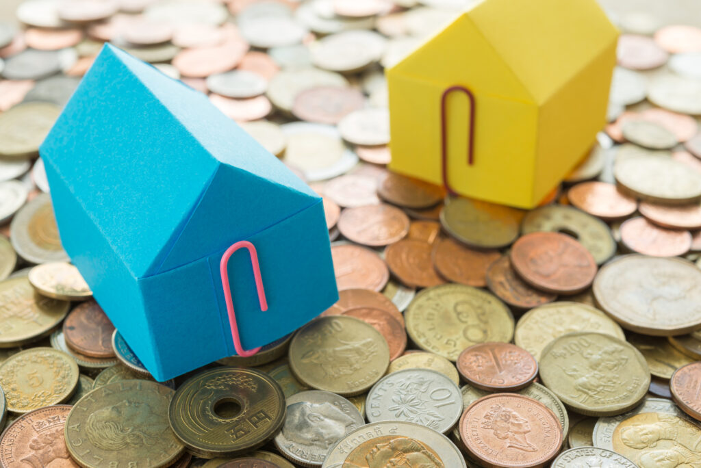 Blue and yellow toy houses with paper clips attached sitting on a pile of money.