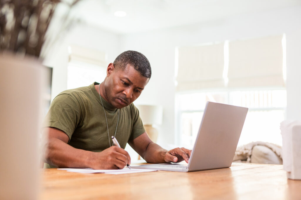 Man in military-style shirt comparing education benefits, working at a laptop and taking notes.