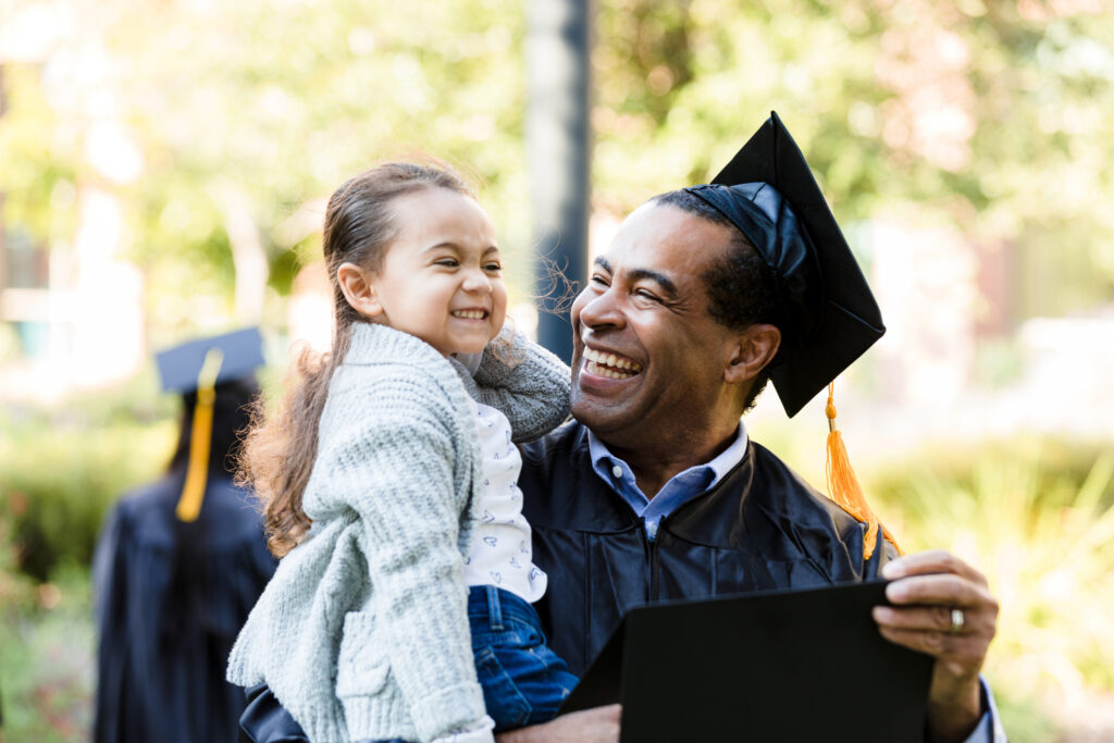 Smiling graduate in cap and gown holding a young girl, celebrating outdoors on graduation day—joyful moment likely made possible with GI Bill benefits.