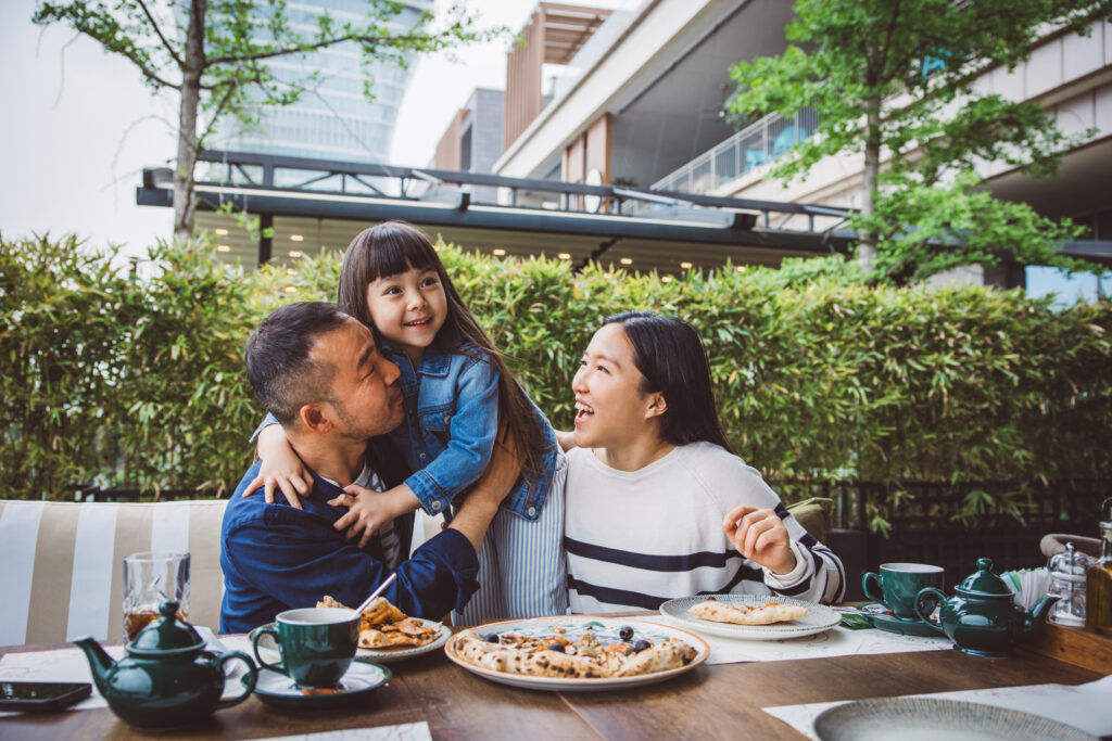 A joyful family of three enjoys an outdoor meal together at a restaurant, smiling and sharing a warm moment—supported by military Basic Allowance for Subsistence (BAS).