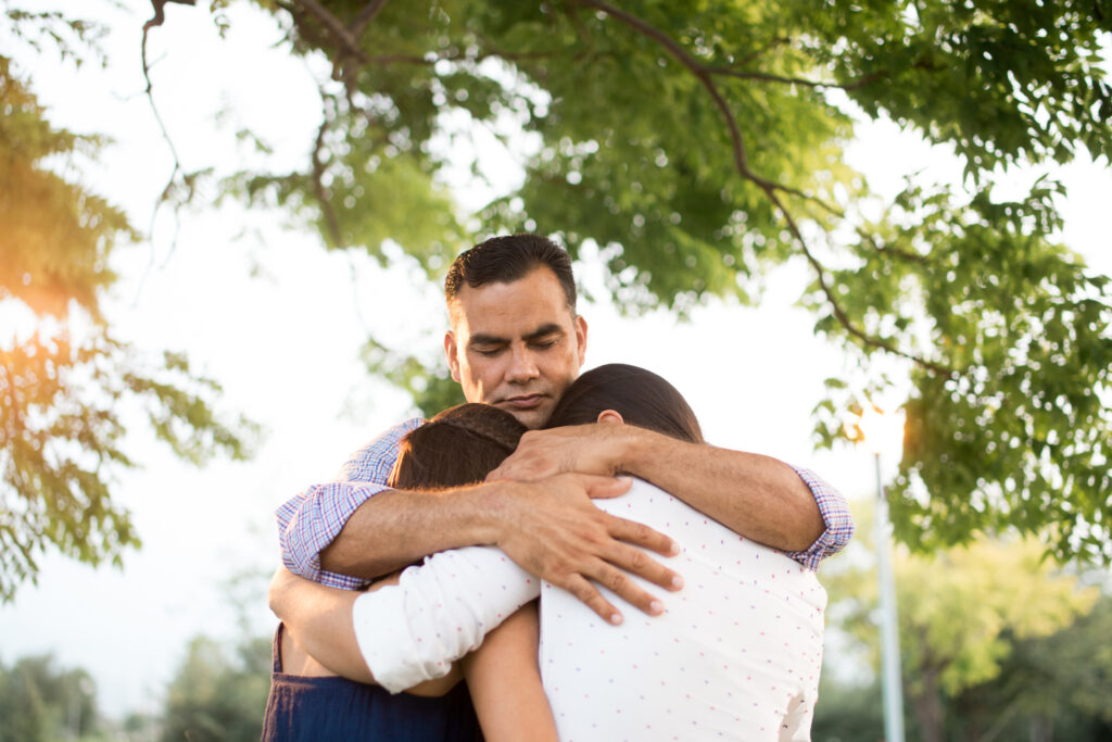 A father embraces his two children outdoors under a tree, eyes closed in a moment of emotional connection before an involuntary family separation.