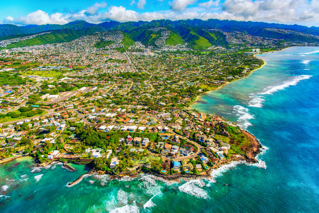 Aerial view of a seaside Hawaiian neighborhood: turquoise ocean waves breaking along a rocky shore, rows of homes hugging the coastline, and lush green mountains rising in the background under a bright blue sky.